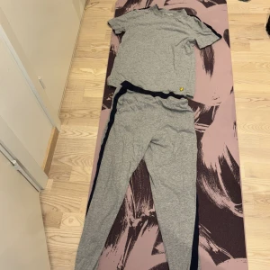 Grå t-shirt från Lyle & Scott och gråa Lyle & Scott byxor - Säljer en grå t-shirt från Lyle & Scott med korta ärmar och rund hals. Tröjan har en svart rand längs axeln och ett gult logomärke på bröstet. Tillverkad i mjuk bomull, perfekt för en chill och sportig stil.