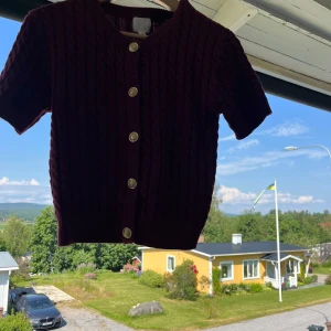 Vinröd kabelstickad kofta med kort ärm - Vinröd kabelstickad kofta med korta ärmar och snygga guldfärgade knappar framtill. Tröjan har rund halsringning och ribbade muddar. Perfekt för dig som vill ha en klassisk men trendig look. Använd fåtal gånge så väldigt bra skick❤️