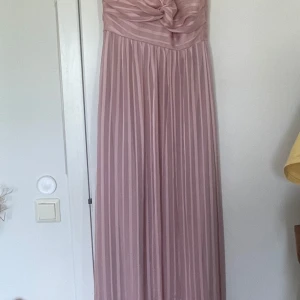 Ljusrosa långklänning från Esprit - Superfin ljusrosa långklänning från Esprit med axelbandslös design och veckad överdel. Kjolen är har subtila ränder och tyget har en mjuk, glansig känsla. Perfekt för bal eller student! Säljes med originaltagg kvar.