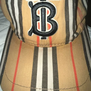 Randig keps från Burberry i bomull - Snygg keps från Burberry med klassiskt randigt mönster i beige, svart, vitt och rött. Framtill finns en broderad TB-logga i svart och vitt. Kepsen är tillverkad i 100% bomull och har justerbar rem baktill för perfekt passform. Ser snyggare i verkligheten!😍