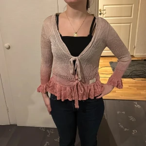 Odd Molly kofta med volang och knyt - Supersöt kofta från Odd Molly i ljusrosa och beige toner. Den är stickad i bomull, har trekvartsärm, volangkant nertill och vid ärmslut samt knyt framtill. Perfekt för dig som gillar romantiska detaljer och en feminin look.