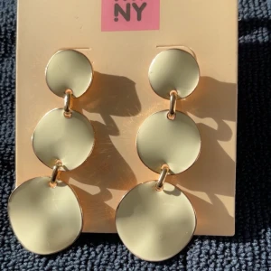 Guldiga statement-örhängen NY - Snygga guldiga statement-örhängen från NY med tre stora, runda plattor i rad. De har en cool, modern look och glänser superfint i ljuset. Perfekta för att lyfta vilken outfit som helst och ge en trendig touch.