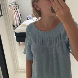 Ljusblå klänning från Daisy Grace - Superfin ljusblå klänning från Daisy Grace i storlek M. Klänningen har en lös passform, kort ärm och ett snyggt rynkat tyg med bred rundad halsringning. Perfekt för varma dagar och enkel att styla med sneakers eller sandaler.