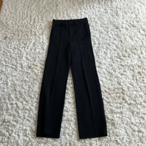 Svarta kostymbyxor från Kappahl - Stilrena svarta kostymbyxor från Kappahl i storlek 140. Byxorna har raka ben, pressveck framtill, hällor för bälte och sidfickor. Perfekta för en dressad look och passar till skjorta eller t-shirt.