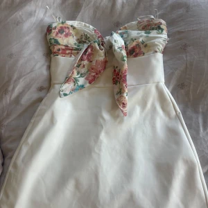 Vit kort klänning med blommig rosett Zara - Superfin vit kort klänning från Zara med blommigt mönster upptill och stor rosett framtill. Axelbandslös design och figurnära passform. Perfekt för sommarens alla tillfällen. Materialet känns mjukt och lätt, och blommorna går i rosa, grönt och blått.
