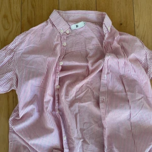 Randig rosa skjorta från jp 1999 - Säljer en klassisk skjorta från jp 1999 i storlek S. Skjortan är rosa och vit med smala ränder, har knappar hela vägen och en snygg krage. Perfekt för dig som gillar en stilren och fräsch look. Materialet känns lätt och passar bra till många outfits.