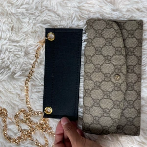 Gucci axelväska med kedja och GG-mönster - Snygg axelväska från Gucci med klassiskt GG-mönster i beige och brunt. Väskan har guldfärgad kedja och tryckknapp framtill. Insidan är fodrad i turkost skinn med flera kortfack. Perfekt för dig som vill ha en stilren och lyxig accessoar.