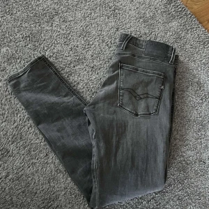Replay Anbass grå jeans, stl 33 - Snygga grå jeans från Replay, modell Anbass. Klassisk femficksdesign med raka ben och diskreta slitningar. Jeansen har en mörkgrå tvätt och är tillverkade i mjukt denimtyg som sitter skönt hela dagen.