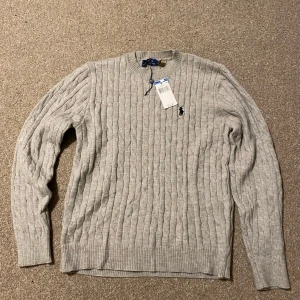Grå kabelstickad tröja från Polo Ralph Lauren - Kabelstickad grå tröja från Polo Ralph Lauren med klassisk svart logga broderad på bröstet. Tröjan har rund halsringning, ribbade muddar och är långärmad. Perfekt för dig som gillar stilren och tidlös design.
