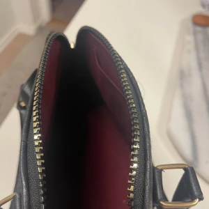 svart handväska i skinn - Säljer en svart handväska med klassiskt monogrammönster i skinn. Väskan har guldfärgade metalldetaljer, dubbla handtag och dragkedja upptill. Insidan är fodrad i vinrött tyg och botten har skyddande nitar. Perfekt för dig som gillar lyxiga accessoarer.