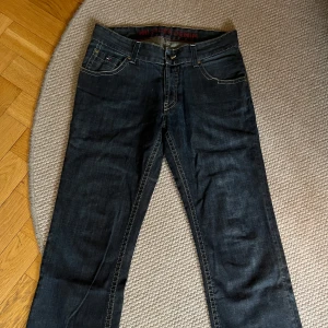 Mörkblå Hilfiger Denim jeans - Snygga mörkblå jeans från Hilfiger Denim med klassisk femficksdesign och kontrastsömmar. Jeansen har raka ben och subtila detaljer som läderpatch bak och logga på fickan. 