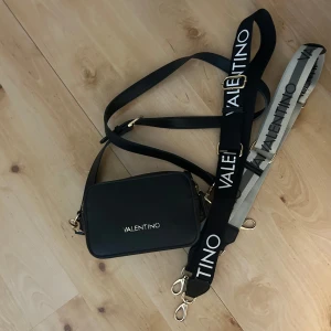 Svart axelväska från Valentino - Snygg svart axelväska från Valentino med guldiga detaljer och två breda axelband, ett svart och ett grått, båda med tydlig logotyp. Väskan är rektangulär och har dragkedja upptill. Perfekt för att lyfta vilken outfit som helst.