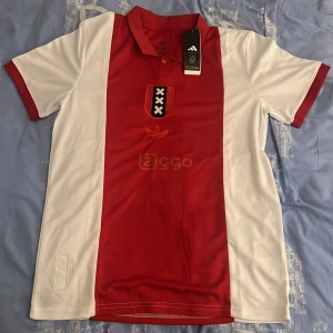 Ajax fotbollströja Adidas röd/vit - Ajax hemmatröja från Adidas med klassisk röd mittpanel och vita sidor. Tröjan har korta ärmar, krage med knappar och Ajax-emblem samt Ziggo-logga på bröstet. Tillverkad i lätt och ventilerande polyester, perfekt för match eller träning.