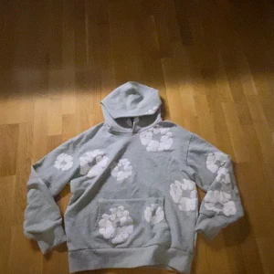 Denim Tears hoodie - Säljer en grå hoodie från Denim Tears med stora vita blommor tryckta över hela tröjan. Hoodien har huva, känguruficka och långa ärmar. Perfekt för dig som gillar streetwear och vill sticka ut med en unik design.