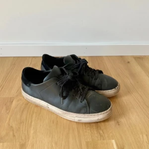 Sneakers från Axel Arigato - Stilrena svarta sneakers från Axel Arigato med ovandel i skinn och svart mockadetalj vid hälen. Klassisk rund tå, svarta skosnören och vit sula med logga baktill. Perfekta för en clean och modern look. Välanvända. 