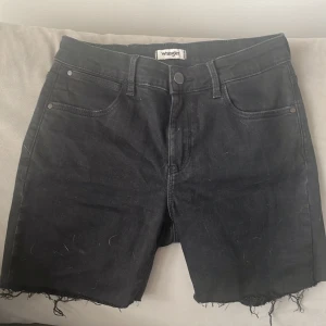Svarta jeansshorts från Wrangler - Svarta jeansshorts från Wrangler med råa, fransiga benslut och klassisk femficksdesign. Modellen har hög midja och raka ben, perfekt för en chill och avslappnad look. Tidlös svart färg som passar till allt och robust denimkänsla.