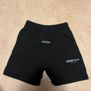 Svarta shorts Essentials Fear of God - Svarta shorts från Essentials Fear of God med elastisk midja och snörning. Klassisk, enkel design med logga framtill och en bakficka. Perfekta för chill dagar eller streetwear-stil. Mjukt material som känns skönt mot huden.