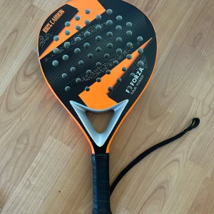 Padelrack  - Bilderna visar ett padelracket av märket FZ Forza, modell Tour Power, tillverkat i 100% kolfiber. Racketet har tydliga tecken på användning med repor och märken längs kanten. Perfekt för dig som vill ta ditt padelspel till nästa nivå!