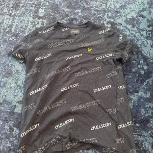 Svart Lyle & Scott t-shirt med logga - Svart t-shirt från Lyle & Scott med vit logotyp tryckt över hela tröjan och den klassiska gula fågeln broderad på bröstet. T-shirten har rund hals, korta ärmar och är gjord i mjuk bomull. Perfekt för en chill och stilren look.