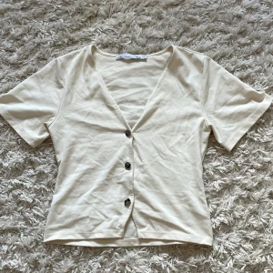 Beige kortärmad blus med knappar - Superfin beige blus från 157 med korta ärmar och djup v-ringning. Toppen har tre unika, silvriga knappar framtill som ger en cool touch. Tillverkad i mjuk bomull, perfekt för en chill och stilren look. Helt oanvänd.
