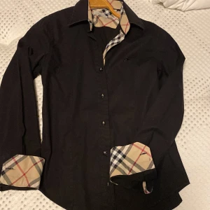 Svart Burberry skjorta med rutiga detaljer - Säljer en svart skjorta från Burberry med klassiska rutiga detaljer på insidan av kragen och manschetterna. Skjortan har lång ärm, knappar framtill och är i bomull. Perfekt för dig som vill ha en stilren look med en touch av lyx. För liten för mig som är 171 skulle säga att den passar 163 och under beroende på hur mycket du väger