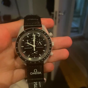 Omega x Swatch MoonSwatch svart klocka - Snygg Omega x Swatch MoonSwatch med svart urtavla och svart textilband. Klockan har kronograf, tachymeter och tydliga vita visare. Boetten är i biokeramik och bandet har Omega-logga. Perfekt för dig som gillar klassisk och sportig stil.