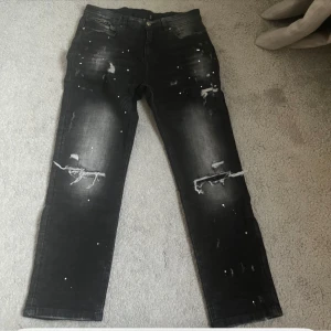 Svarta distressed jeans Supply & Demand - Säljer ett par svarta jeans från Supply & Demand med slitna detaljer och hål på benen. Jeansen har en cool, tvättad look med vita färgstänk och raka ben. Perfekta för dig som gillar streetstyle och vill sticka ut lite extra.