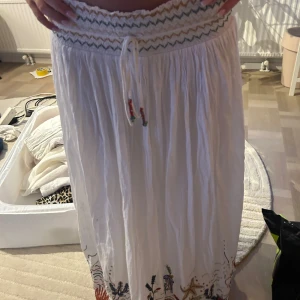 Vit långkjol med broderier och snörning - Superfin vit långkjol med broderade detaljer i färg längs nederkanten och  midja med snörning💘