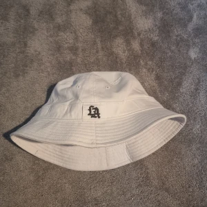 Vit bucket hat med LA-brodyr - Vit bucket hat i bomull från New Yorker med svart LA-brodyr framtill. Klassisk form med bred, nedåtlutande brätte och sydda detaljer runt om. Perfekt för dig som vill ha en clean och stilren look med streetkänsla.