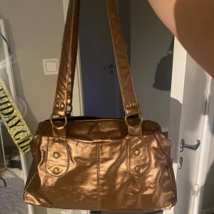 Guldig axelväska med metallic-look - Säljer en snygg axelväska i metallic guld med dubbla axelband och coola nitar. Väskan har dragkedja upptill, flera innerfack och ljusgul insida. Dragkedjorna upptill och på baksidan är trasiga men syns inte när man bär den💕 