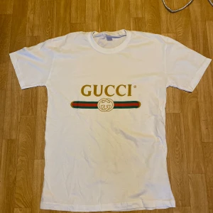 Gucci T Shirt S/M - Vit t-shirt från Gucci med klassisk logga i guld, rött och grönt tryck på bröstet. T-shirten har rund hals och korta ärmar, tillverkad i mjuk bomull. Perfekt för dig som vill ha en clean och lyxig vibe.