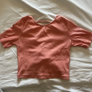 Aprikos topp från monki - Aprikosfärgad ribbad topp med korta ärmar och snörningsdetalj i ryggen från monki. Toppen har en croppad passform. Toppen är välanvänd men fortfarande i bra skick. Det finns en liten noppa/minireva framtill (se bild), knappt märkbar vid användning. Toppen ser lite mörkare ut på bilderna än vad den är i verkligheten, fler bilder kan fås vid intresse💕