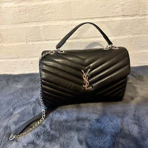Svart quiltad YSL handväska i skinn - Säljer en svart quiltad handväska från YSL med silverfärgad kedja och ikonisk YSL-logga framtill. Väskan är tillverkad i mjukt skinn och har både handtag och axelrem i kedja. Snygg och stilren design med chevronmönster som ger en lyxig känsla.