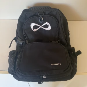 Svart ryggsäck från Nfinity - Säljer en svart ryggsäck från Nfinity med stort fack, flera dragkedjor och meshfickor på sidorna. Ikonisk logga framtill och praktiskt handtag upptill. Perfekt för att bära allt du behöver till träning eller skola.
