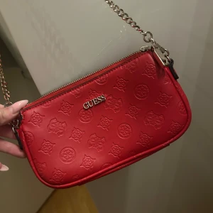 Röd axelväska från Guess med kedja - Snygg röd axelväska från Guess med silverfärgad kedjerem och präglat logomönster över hela väskan. Väskan har dragkedja upptill och en stilren, kompakt form som passar perfekt till din outfit.