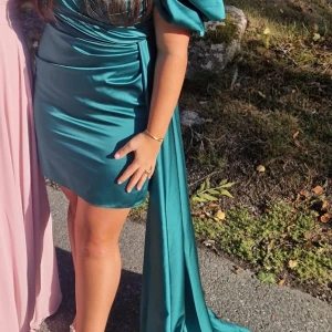 Siden klänning - Snygg  kortklänning i satin med markerad midja, puffiga korta ärmar och en lång draperad detalj på sidan. Klänningen har en elegant omlottkänsla och passar perfekt för dig som vill sticka ut.