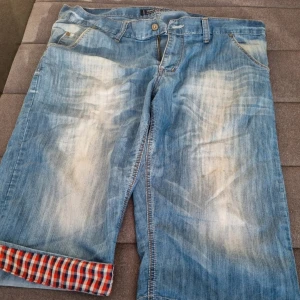 Tommy Hilfiger jeansshorts blå - Snygga jeansshorts från Tommy Hilfiger i ljusblå denim med slitna detaljer och klassiska fickor. Insidan av bensluten har ett rutigt tyg i rött, blått och vitt som syns om man viker upp dem. Perfekta för en chill sommarstil.