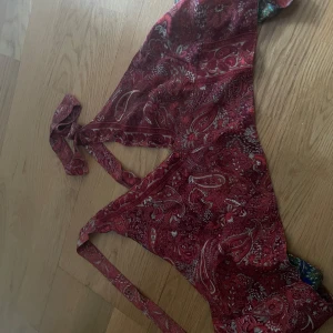 Färgstark omlottklänning med paisleymönster - Säljer en unik omlotttopp i paisleymönster med röda och blå partier. toppen har knytband i midjan och är ärmlös, perfekt för dig som gillar bohemisk stil och vill sticka ut. Materialet känns lätt och svalt, och modellen är kort.