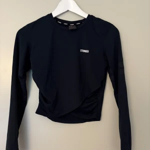 Svart långärmad topp från Stronger - Svart långärmad topp från Stronger med diskret logga på bröstet. Snyggt omlott-draperad framtill och tight passform som framhäver figuren. Perfekt för träning eller en sportig look. Tillverkad i stretchigt material för maximal komfort.