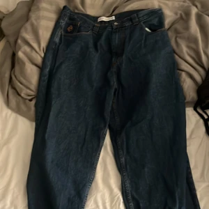 Big Boy blå baggy jeans - Säljer ett par mörkblå baggy jeans från Big Boy med orangea kontrastsömmar och broderad smiley på myntfickan. Jeansen har klassisk femficksdesign, bred passform och en blå patch med logga bak i midjan. Perfekta för dig som gillar streetwear vibes.