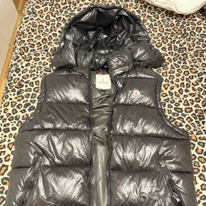 Svart dunväst från Moncler med huva - Säljer en svart, glansig dunväst från Moncler med avtagbar huva och dragkedja framtill. Västen har två fickor med dragkedja och klassisk Moncler-logga på bröstet. Perfekt för lager-på-lager och riktigt snygg till streetwear.säljs pga den e lite för stor för mig helt ny oanvänd