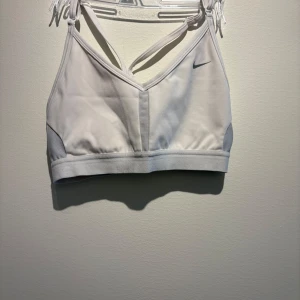 Vit sporttopp från Nike Dri-Fit S - Sportig vit linnetopp från Nike med Dri-Fit-teknologi. Snyggt korsade axelband i ryggen och meshpaneler på sidorna för extra ventilation. Perfekt för träning och aktiva dagar. Enkel design med diskret Nike-logga framtill.