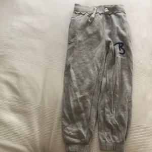 Grå mjukisbyxor från Bondelid - Grå mjukisbyxor från Bondelid med broderad blå B-logga och vit Bondelid-text. Byxorna har resår och snörning i midjan samt muddar vid bensluten. Perfekta för chill dagar eller träning. Tillverkade i mjuk bomullsmix för extra komfort.