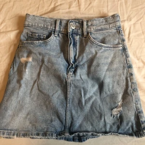 Ljusblå jeanskjol från H&M  - Säljer en ljusblå jeanskjol från H&M med klassisk femficksdesign och slitna detaljer framtill. Kjolen har bälteshällor, knapp och dragkedja framtill samt raka bakfickor. Perfekt för en chill och avslappnad look.