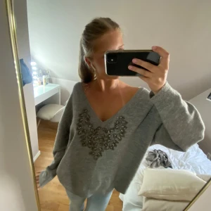 Grå stickad tröja med pärldetaljer - Supermjuk grå stickad tröja med v-ringning och snygga pärldetaljer framtill. Tröjan har en loose passform och långa ärmar. Använd två gånger🩷