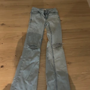 Ljusblå bootcut jeans med broderi - Säljer ett par ljusblå bootcut jeans med slitna knän och råa kanter nertill. Baksidan har coola bakfickor med röd broderad våg. Perfekta för dig som gillar en avslappnad och trendig look.