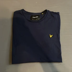 Mörkblå t-shirt från Lyle & Scott - Snygg mörkblå t-shirt från Lyle & Scott med klassisk rund hals och gul broderad logga på bröstet. Enkel och stilren design som passar perfekt till jeans eller shorts. Tillverkad i mjuk bomull för skön känsla hela dagen. Hör av er vid minsta lilla fråga. 