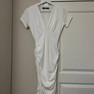 Vit omlottklänning från BikBok XS - Supersnygg vit klänning från BikBok i omlottmodell med v-ringning och korta ärmar. Klänningen har rynkade detaljer längs sidorna som ger en cool draperad effekt. Perfekt för dig som gillar en stilren look med twist.