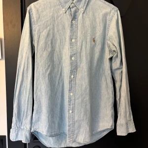 Ljusblå skjorta från Ralph Lauren S - Klassisk ljusblå skjorta från Ralph Lauren i 100% bomull. Skjortan har button-down krage, långa ärmar och knäppning framtill. Ikoniska broderade polospelaren på bröstet ger en stilren och tidlös look. Perfekt för dig som gillar enkel och snygg stil.