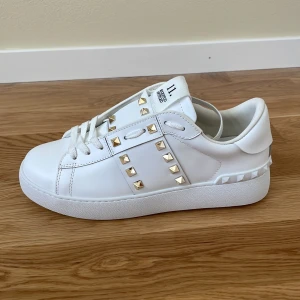 Valentino Rockstud vita sneakers - Snygga vita Valentino Rockstud sneakers i skinn med ikoniska guldiga nitar och chunky sula. Klassisk rund tå, snörning och coola detaljer längs sidorna och hälen. Perfekta för dig som vill sticka ut med en lyxig touch. Säljer då de tyvärr inte passade mig, strl 39 men skulle mer säga att de är som en 38❤️